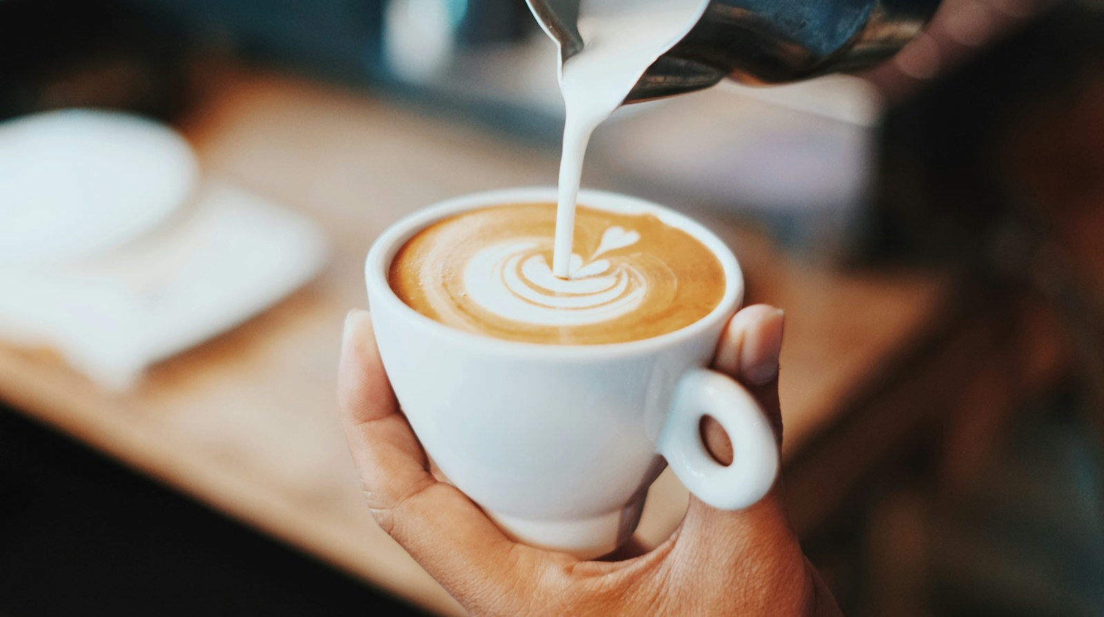 Hands pouring steamed milk into a cup of coffee to create latte art.