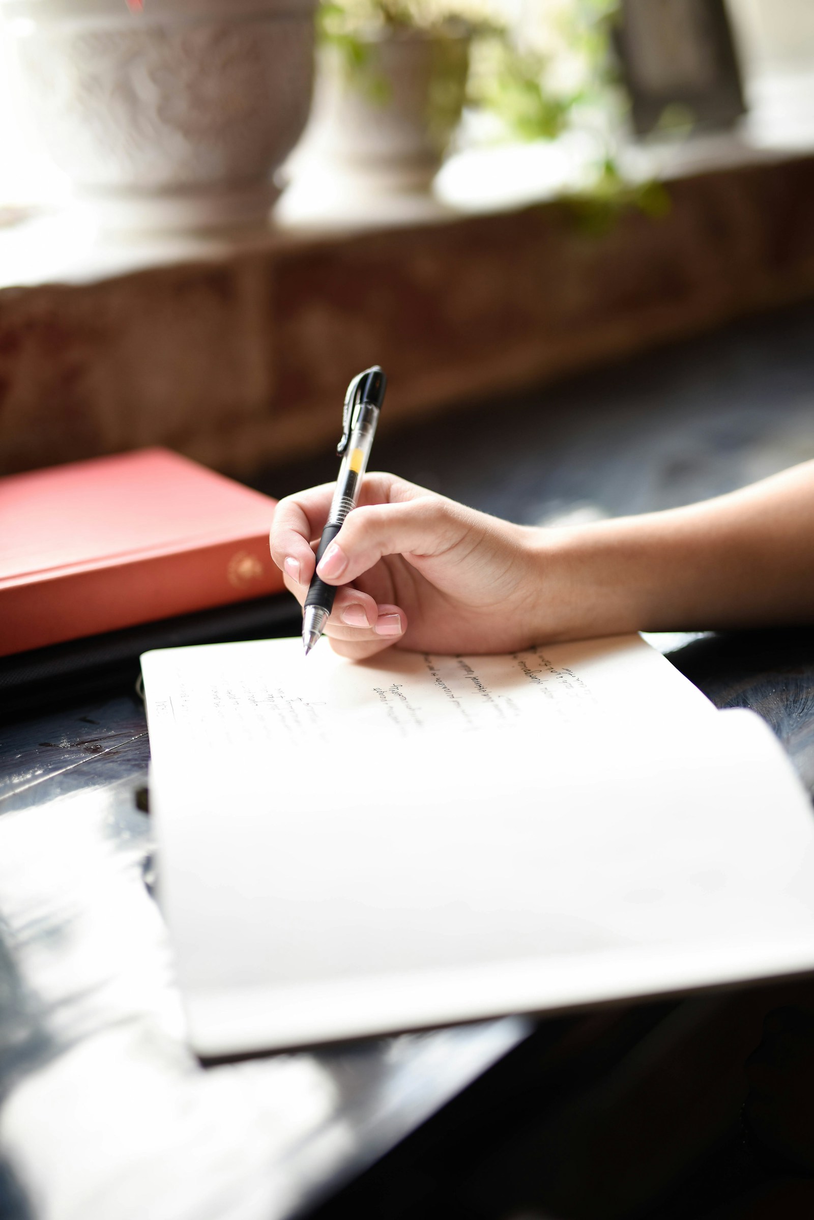 A hand writing in a notebook at a table beside a window.