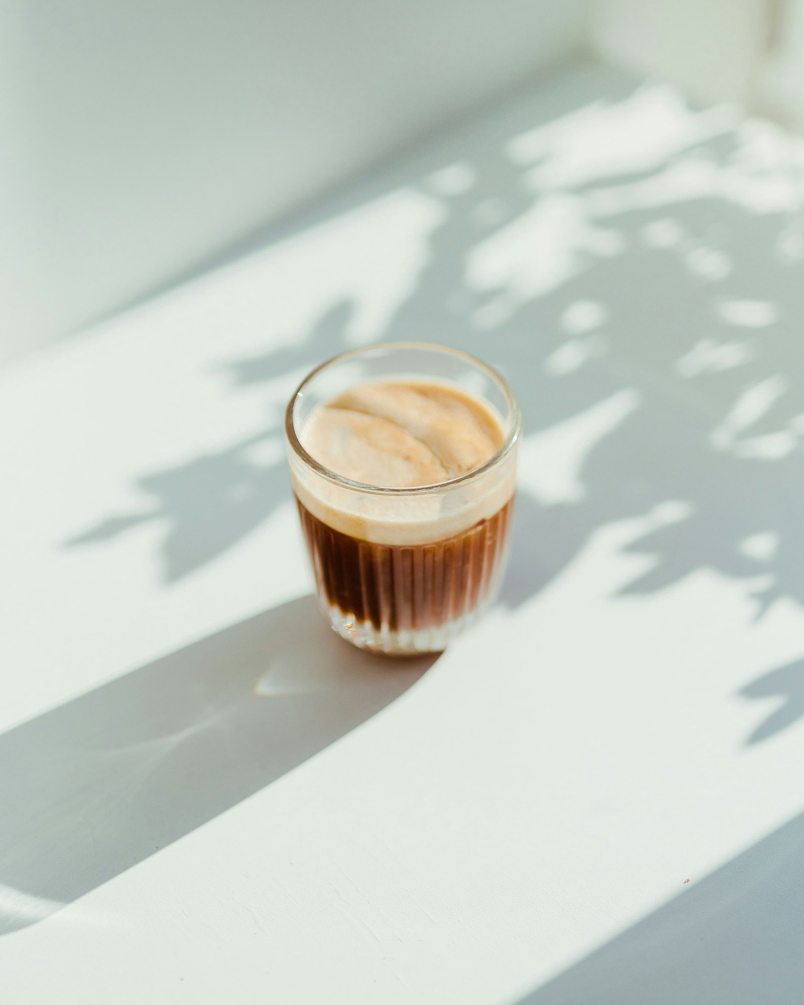 A glass of coffee on a light table crossed by soft leaf shadows.