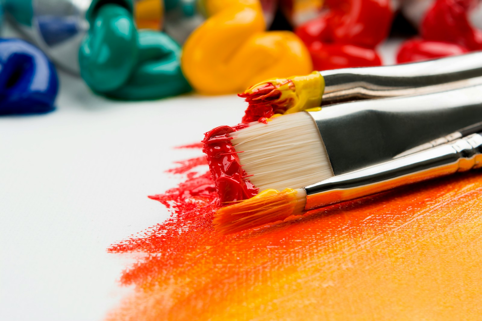 Paintbrushes dragging bright color across a white surface beside paint tubes.