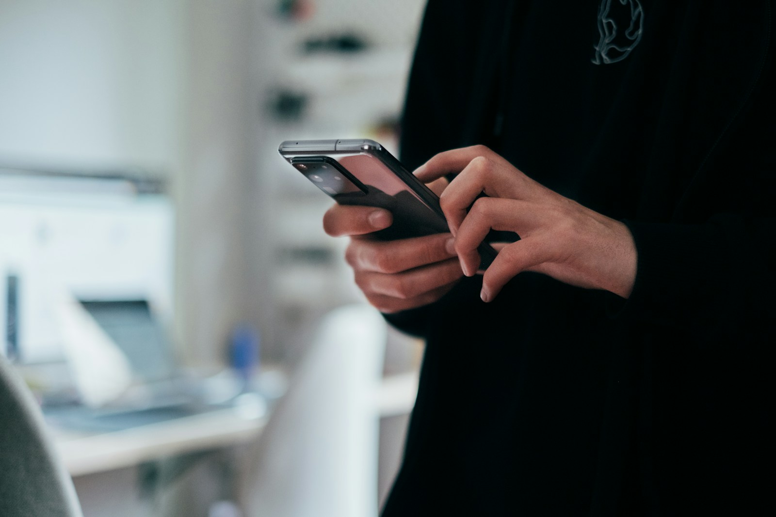 Hands typing on a smartphone in a softly lit work setting.