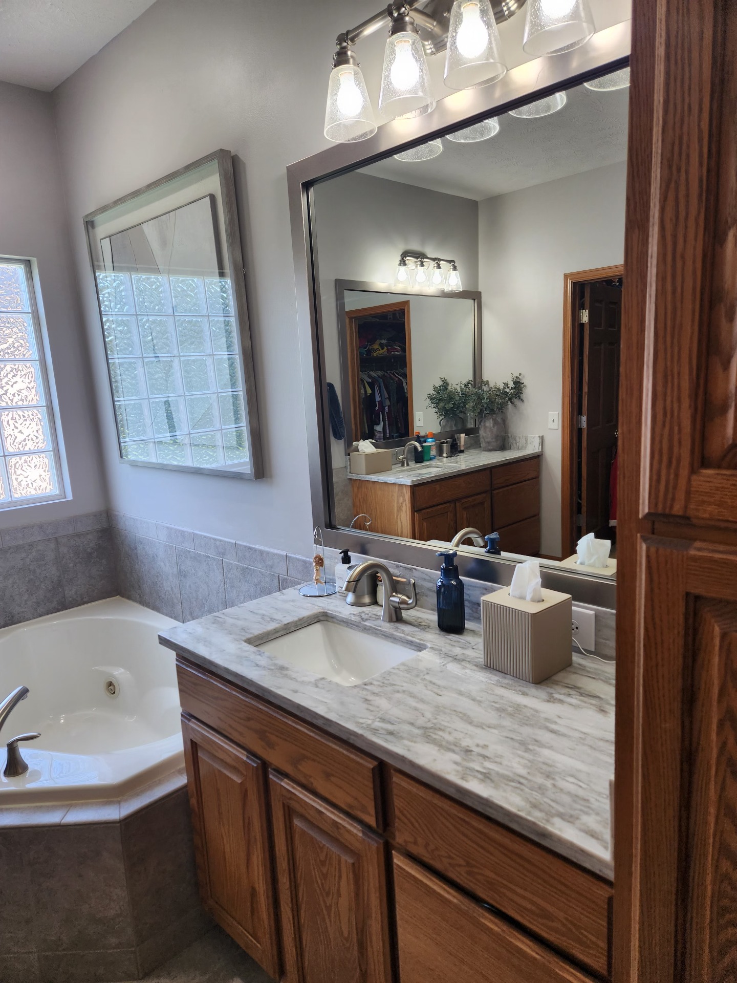 Spacious primary bathroom featuring a marble-look vanity, large framed mirror, warm wood cabinetry, modern lighting, and a corner soaking tub.
